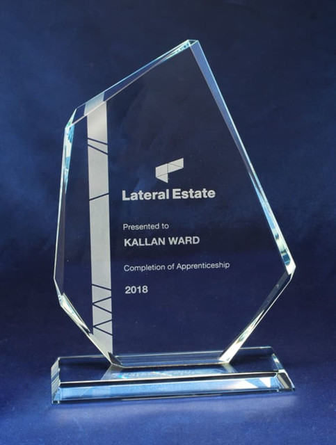 A clear glass award with a unique shape, featuring engraved text and a base, in a blue backdrop. A clear glass award with a unique shape, featuring engraved text and a base, in a blue backdrop.