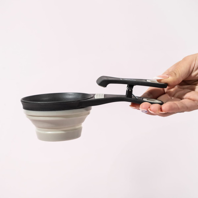 A collapsible silicone pet food scoop and bag clip in black and grey, held by a hand against a light background.