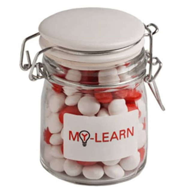 A glass clip lock jar with a white lid, filled with red and white chewy fruit sweets. The jar features a logo. A glass clip lock jar with a white lid, filled with red and white chewy fruit sweets. The jar features a logo.