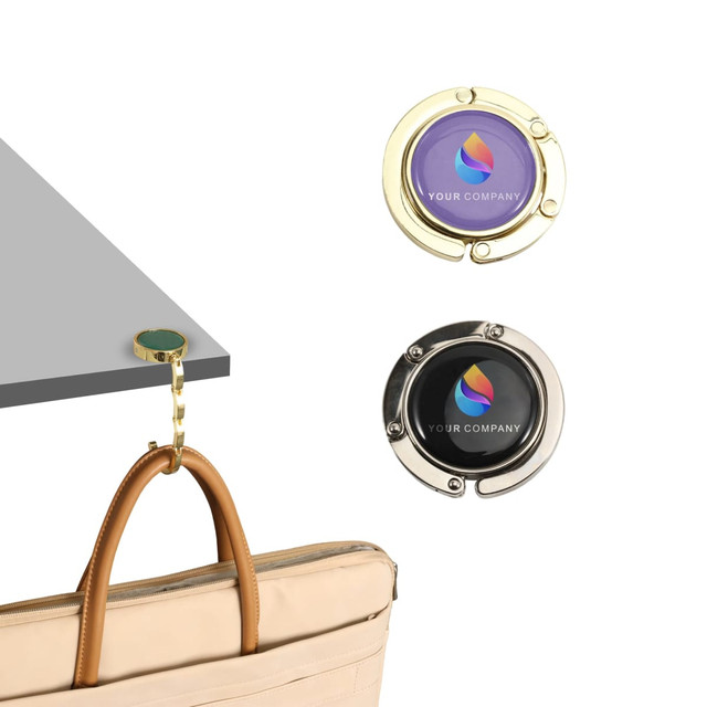 A round foldable desk hook in gold and silver, featuring decorative logos, holding a handbag off a table edge.