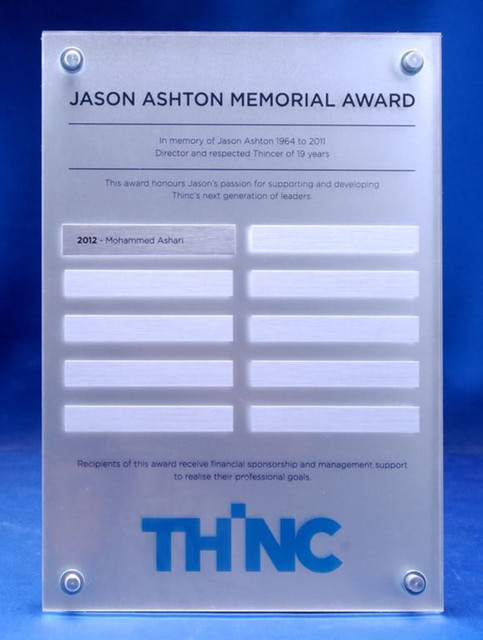 A clear acrylic award plaque with engraved sections for names, featuring a blue logo at the bottom.
