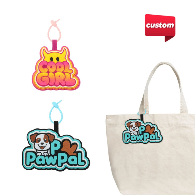 Custom felt hanging tags in pink and blue, featuring playful designs, attached to a beige tote bag.