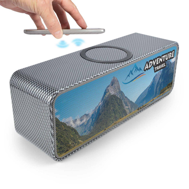 A silver speaker with a scenic mountain design, featuring an inductive charging area on top. It has a logo.