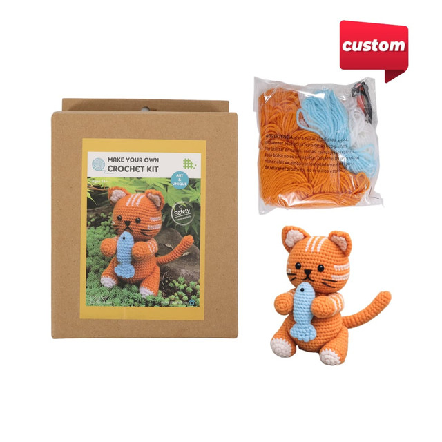 A crochet kit featuring an orange striped kitten with a fish, packaged in a brown box with additional yarn.