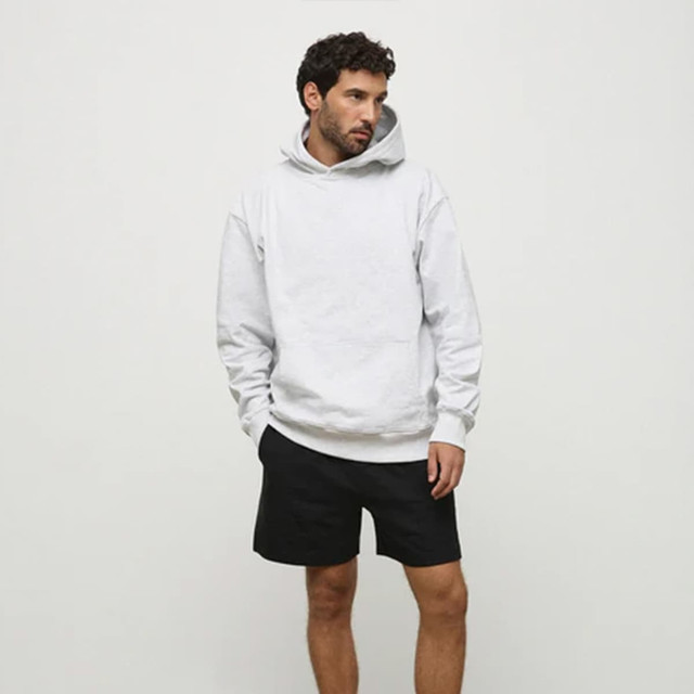 A man wearing a grey hoodie and black shorts stands against a neutral background. The hoodie features a logo.