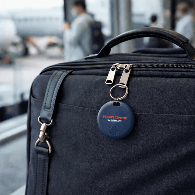 A navy blue tracking device attached to a dark bag, set against a blurred airport background.