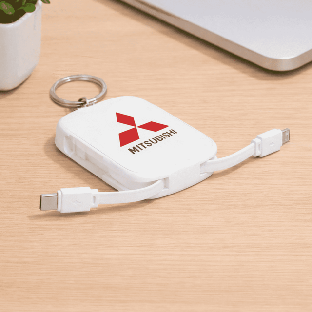 A white power bank keychain with dual charging cables and a logo, resting on a wooden surface next to a laptop.