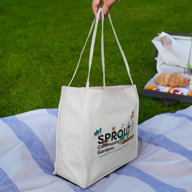 A cream-colored tote bag with two handles, featuring a logo and illustrations, set on a grassy background.