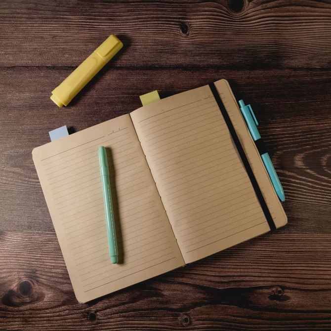 A brown A5 notebook with lined pages, accompanied by a green pen, a blue pen, and a yellow highlighter on a wooden surface.