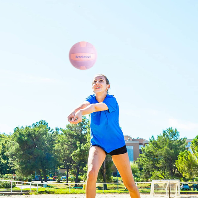 A volleyball is flying through the air as a woman in a blue shirt prepares to hit it outdoors.