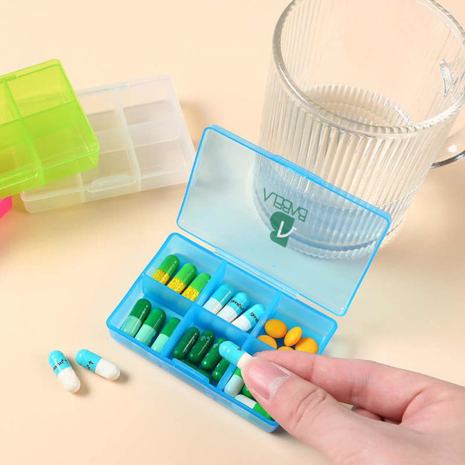 A blue six-compartment pill box is open, filled with various coloured pills, beside a glass of water.