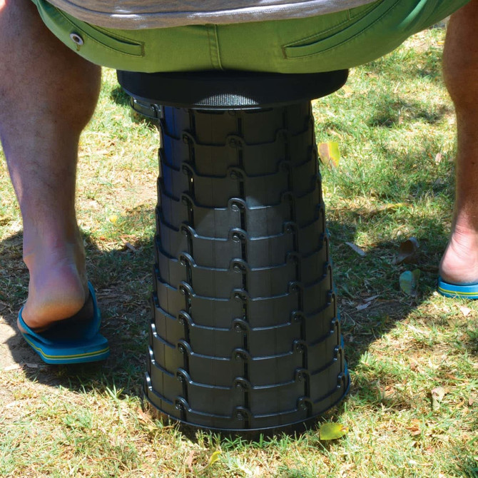 A black telescopic stool sits on grass, with a person in flip-flops sitting above it.