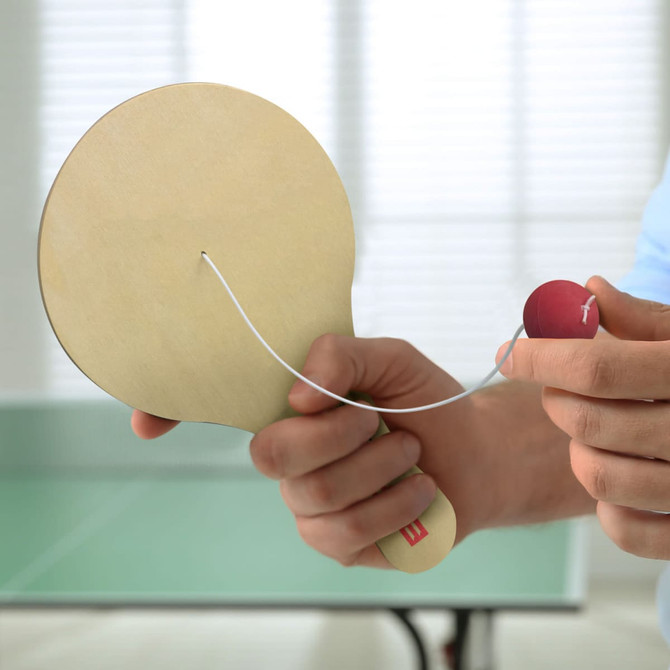 A wooden paddle with a red ball attached by white string, held in a hand against a blurred background.