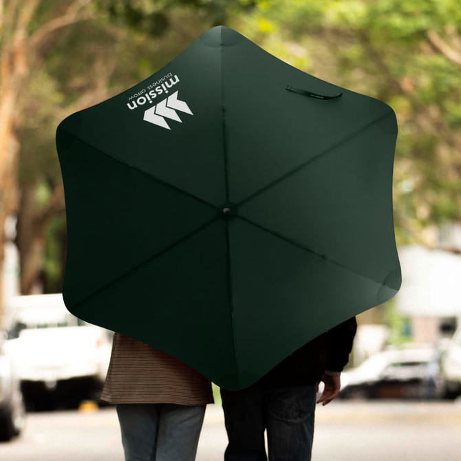 A dark green sport umbrella with a logo, opened and being held by two people, against a blurred outdoor background.