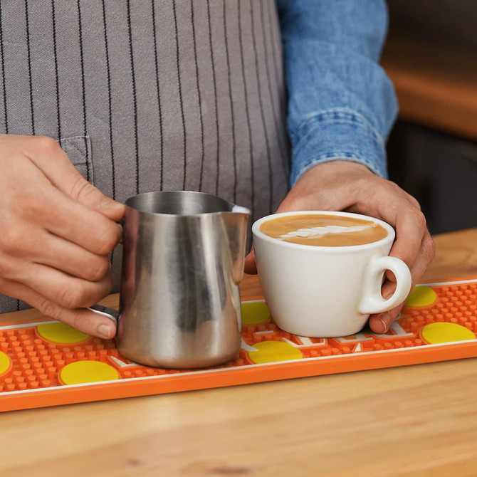 A stainless steel milk jug and a white coffee cup on a vibrant orange PVC bar runner with yellow dots.