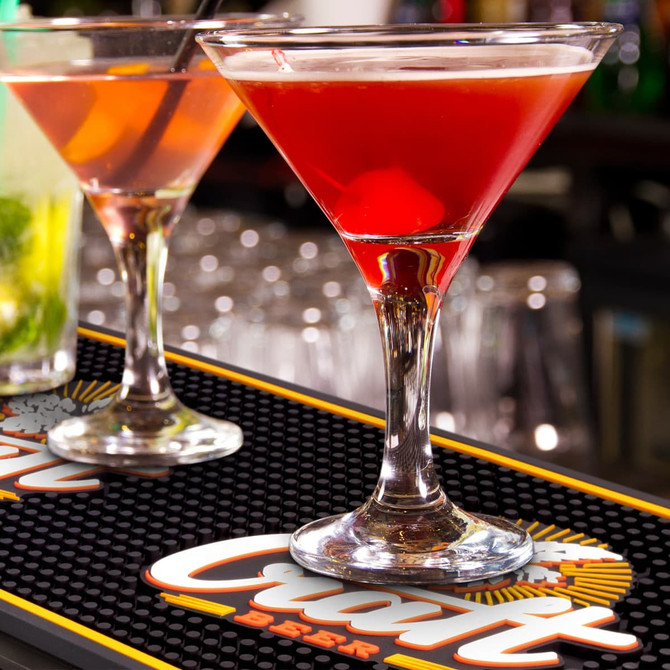 A vibrant red cocktail in a martini glass on a black PVC bar mat, with another drink in the background.