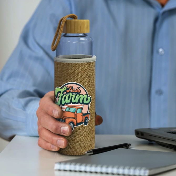 A drink bottle with a clear glass body and natural sleeve, featuring a logo and a bamboo cap.