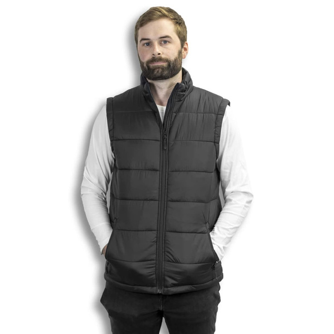 A man in a dark grey puffer vest stands with hands in pockets against a white background, featuring a logo on the vest.
