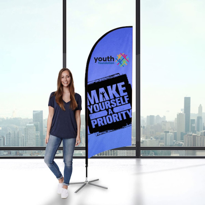 A small feather flag kit in blue with a logo, displayed beside a person in a casual outfit, against a city backdrop.