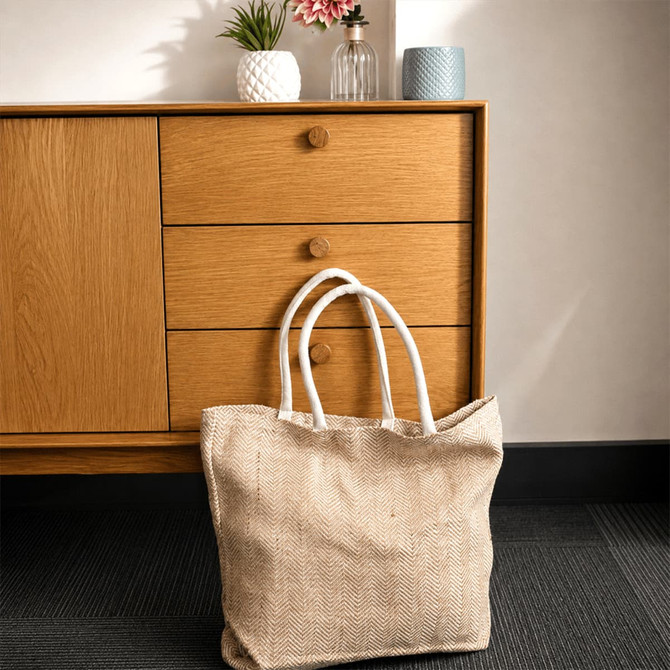 A herringbone jute tote in natural colour with white handles, positioned beside a wooden drawer unit.