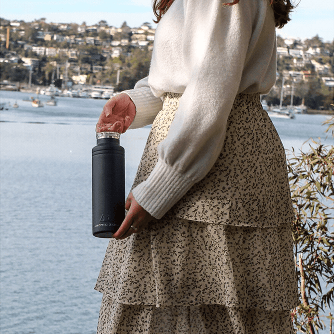 A drink bottle in dark grey with a silver cap, held by a person wearing a white sweater and patterned skirt.