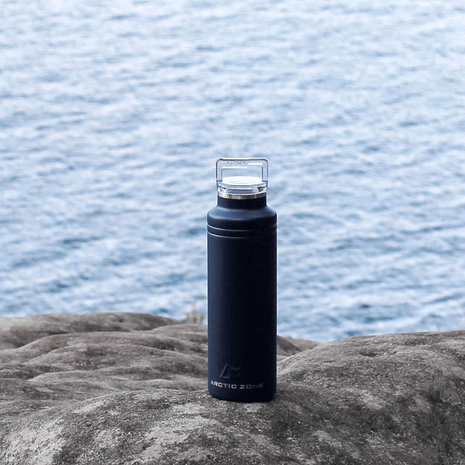 A drink bottle in matte black with a clear lid, featuring a logo, displayed on a rocky surface near water.