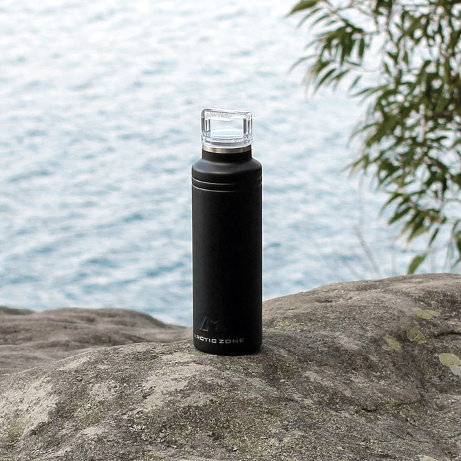 A drink bottle in matte black, featuring a clear cap and a logo, set against a natural rocky background.