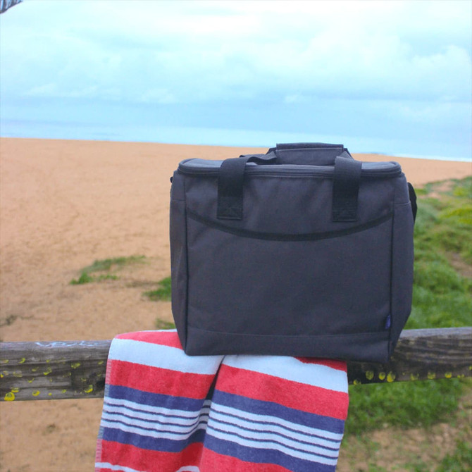A grey cooler bag made from recycled material, placed on a wooden railing beside a beach towel.