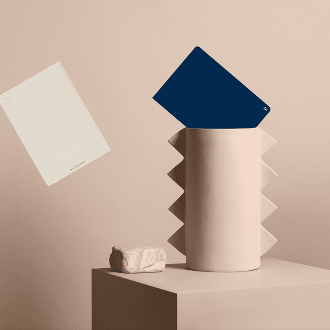 A hardcover notebook with a navy cover and a cream-coloured page, placed next to a textured vase and a stone.