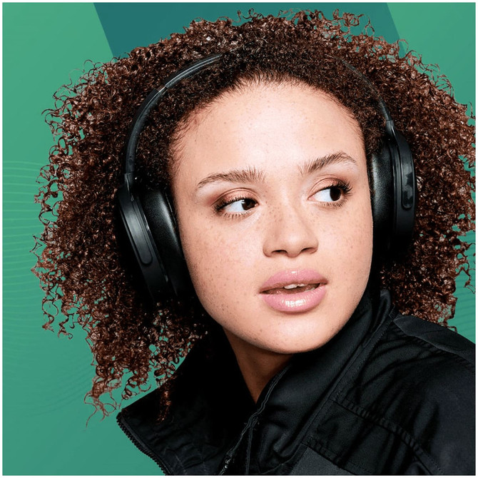 A young woman wearing black Skullcandy Hesh ANC Wireless headphones against a green background.