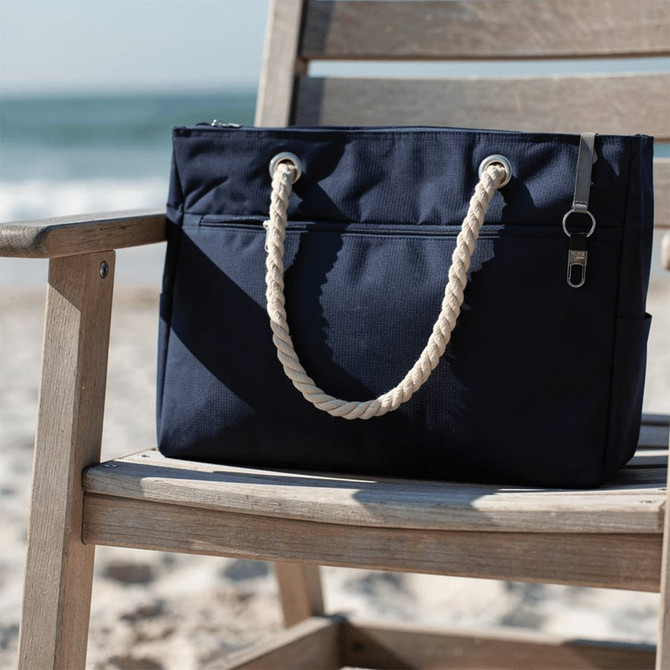 A navy waterproof beach bag with rope handles rests on a wooden chair by the shore. Features a logo.