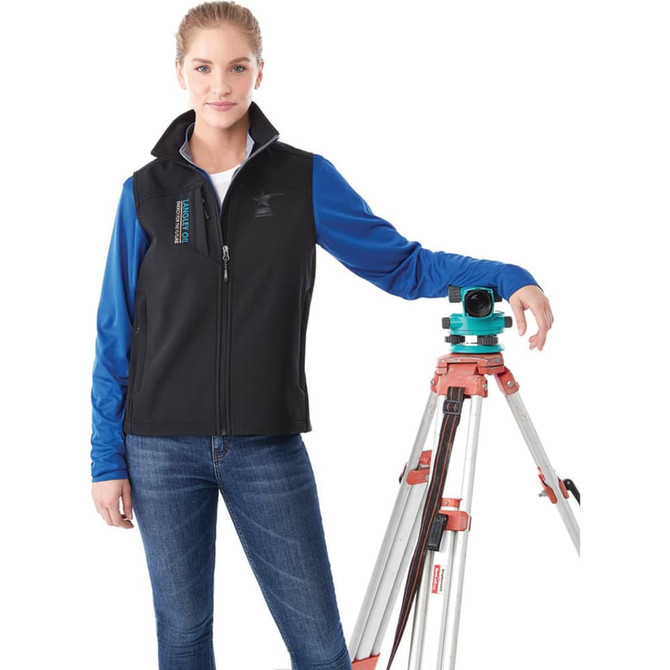 A woman stands beside a tripod, wearing a black and blue Stinson Softshell Vest with a logo on it.