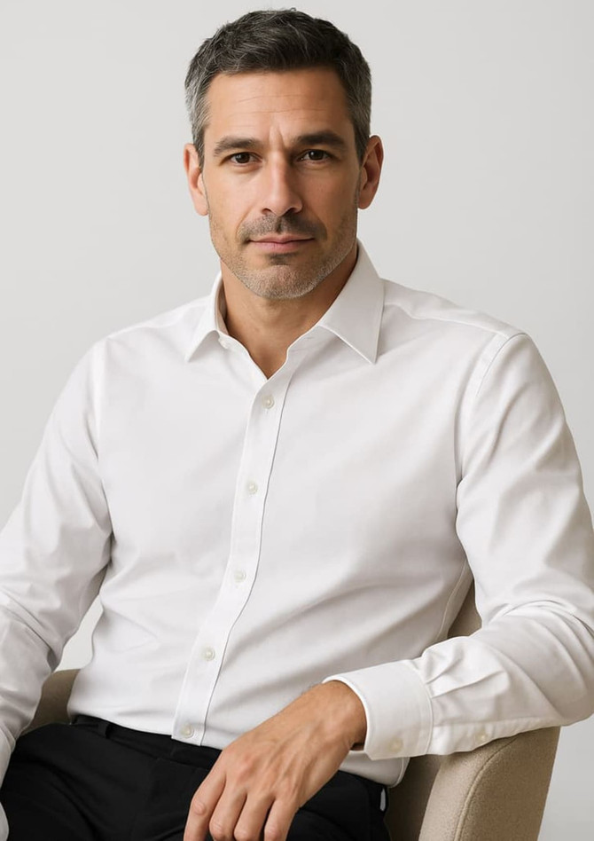 A long sleeve button-up shirt in white, displayed on a seated man. The shirt features a collar and has branding.