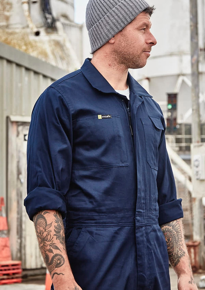 A man wearing a navy blue lightweight cotton drill overall with a logo on the chest, standing in an industrial setting.