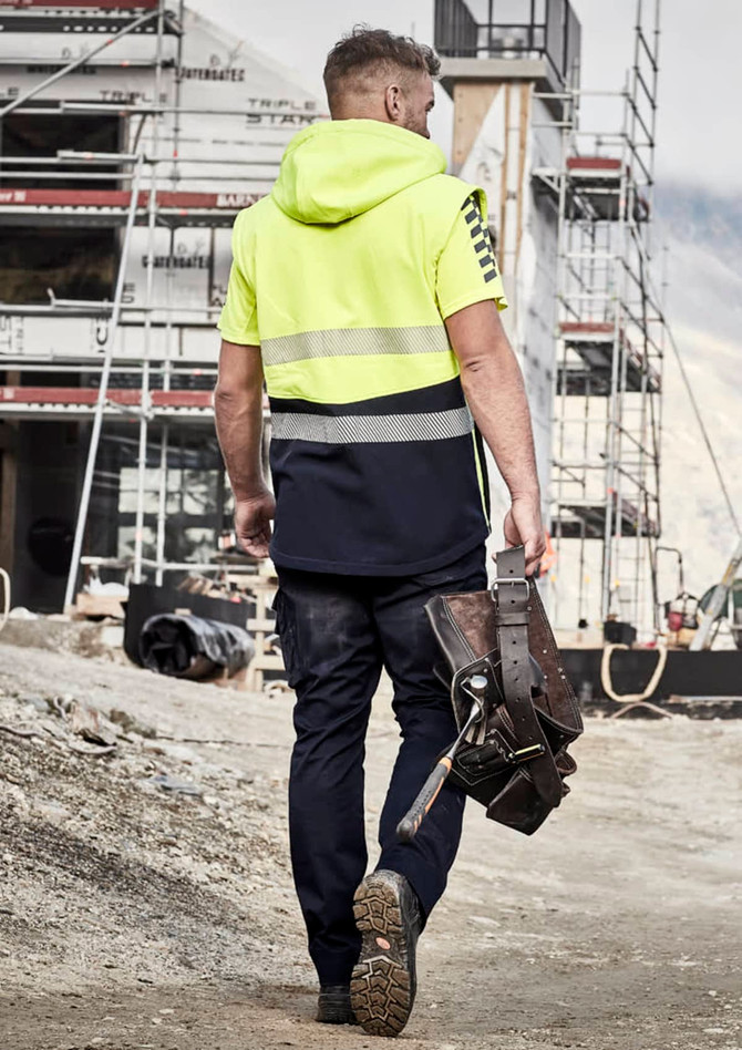 A man in a high-visibility yellow and navy softshell jacket walks at a construction site, holding a tool belt.