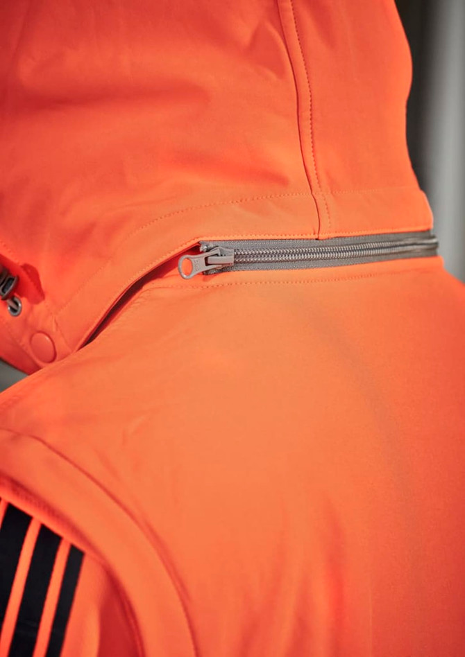 An orange unisex softshell jacket with a zipper and hood, featuring black stripes and a logo.