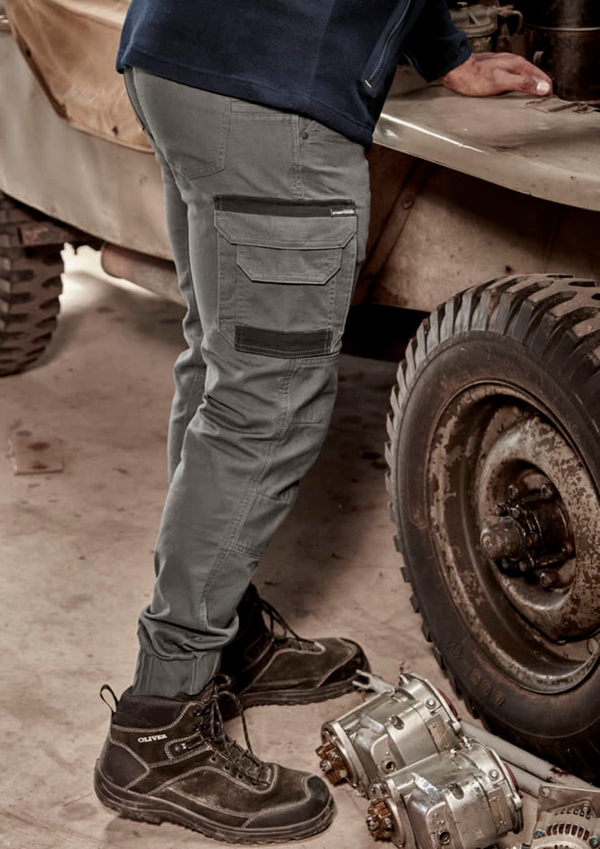 A pair of cuffed heritage work pants in grey, shown with a person standing beside machinery. The pants feature cargo pockets and a logo.