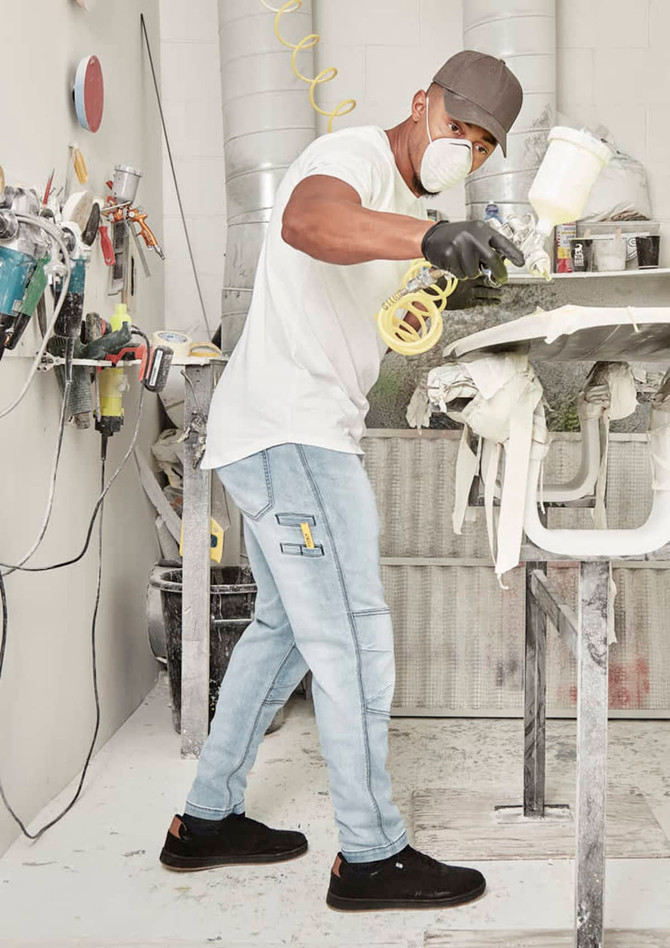 A man wearing light blue stretch jeans and a white t-shirt is spray painting in a workshop.