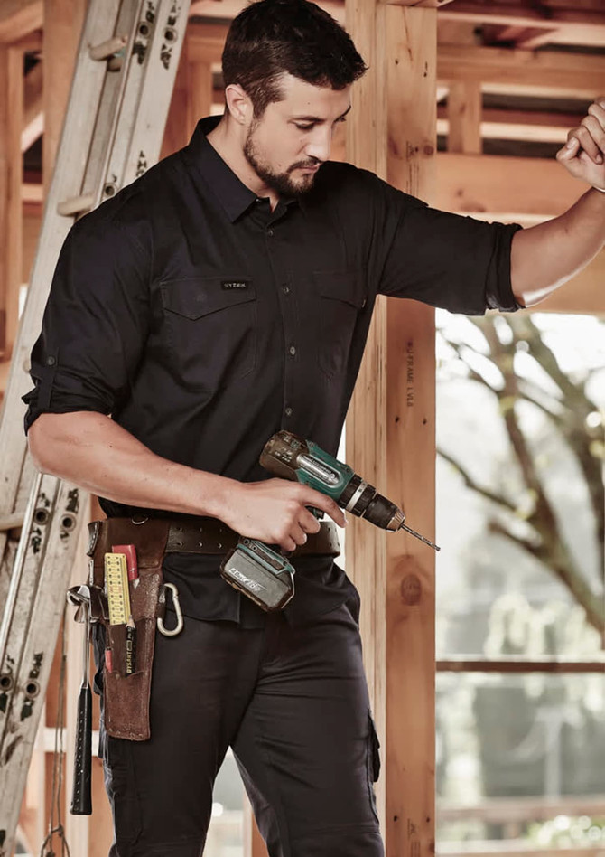 A man in a black rugged long sleeve shirt is holding a power drill, standing inside a construction site.