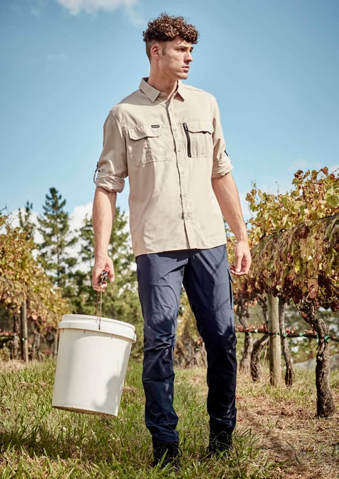 A long-sleeve, button-up shirt in beige, worn with navy pants, against a vineyard backdrop. Features a logo.