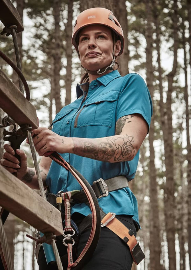 A woman wearing a blue short sleeve outdoor shirt with a harness, set against a forest background.