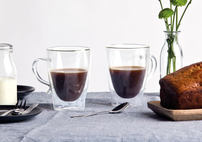 A transparent glass coffee cup set in a neutral setting, with two cups filled with dark coffee and a slice of cake nearby.