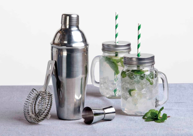 A stainless steel cocktail shaker, a strainer, a jigger, and two glass jars with green straws and mint garnish.