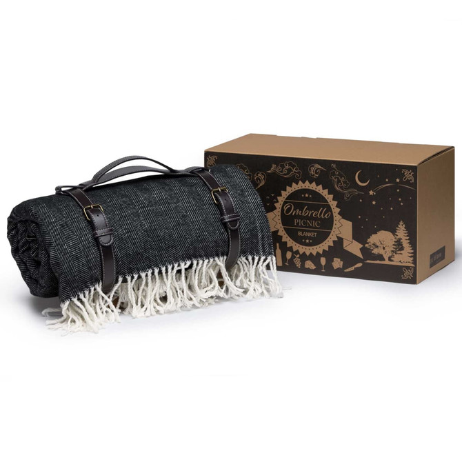 A dark green picnic blanket with a white fringe, folded and secured with straps, beside a brown packaging box.