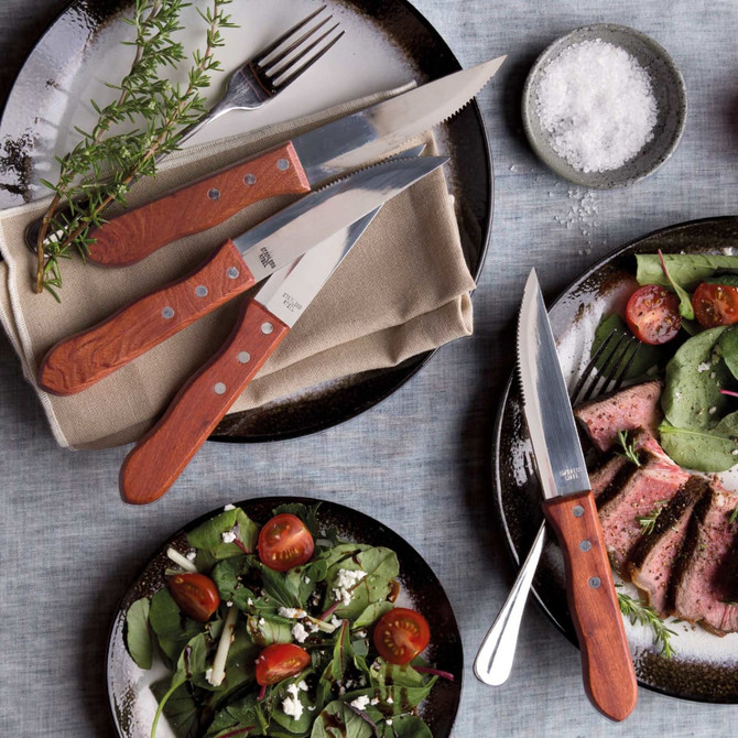 A Steak Knife set featuring six knives with wooden handles, placed on textured plates alongside a fresh salad and steak.