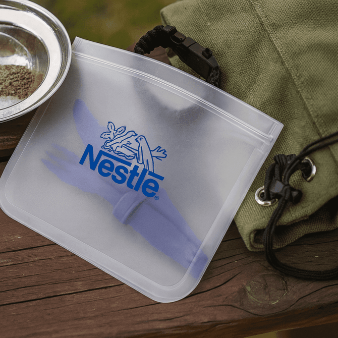 A clear PEVA reusable food storage bag featuring a blue logo, placed on a wooden surface beside a plate and bag.