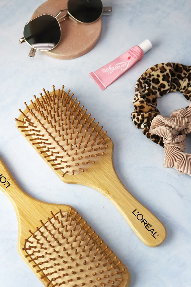 A bamboo hairbrush in a light wooden finish, accompanied by sunglasses, lip balm, and a hair scrunchie.
