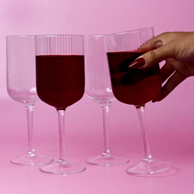 Four clear glass wine glasses with ribbed design, displayed against a pink background, one held with red liquid.