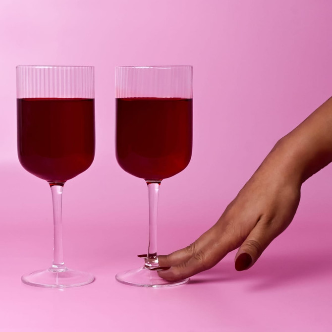 Two clear glass wine glasses with ribbed tops, filled with red liquid, set against a pink background.