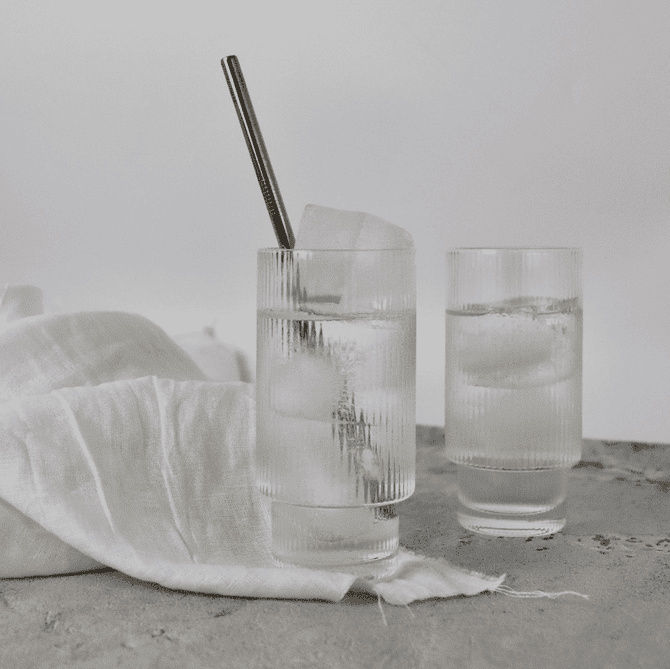 Two ribbed highball glasses filled with clear liquid and ice, alongside a white linen cloth and a metal straw.